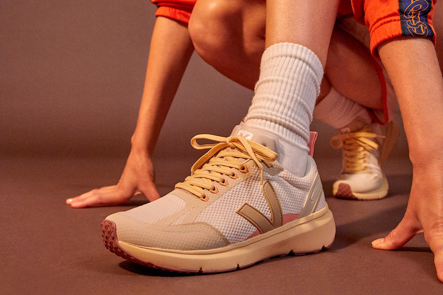 Close-up of a person crouching in beige and pink VEJA Condor sneakers with yellow laces, wearing white socks and orange shorts.