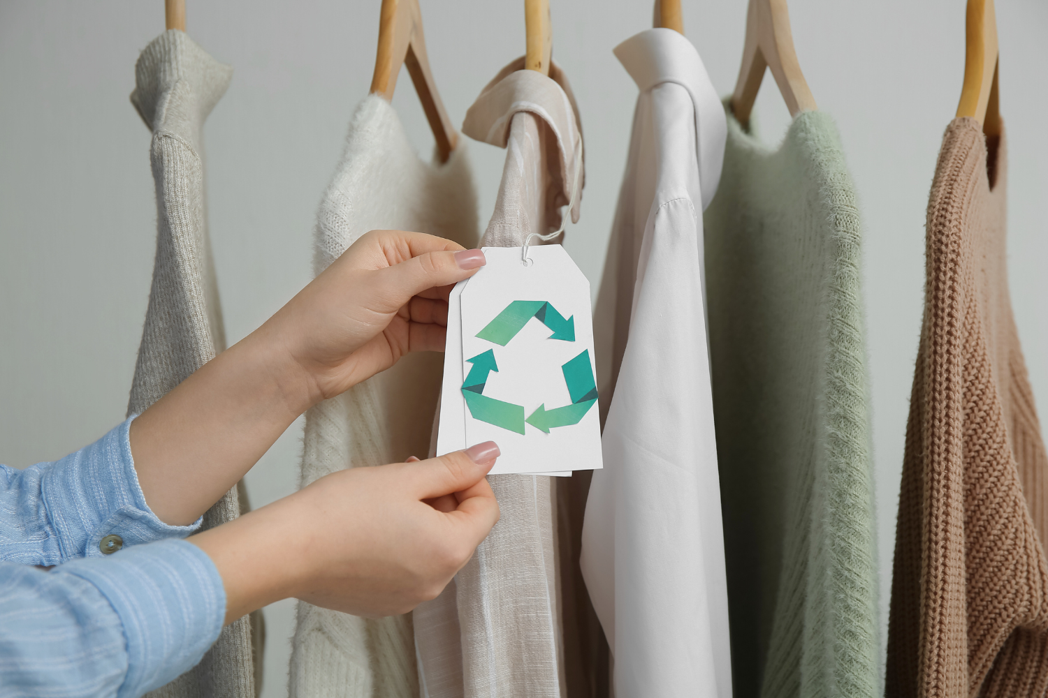 “Hands holding a clothing tag with a green recycling symbol in front of a row of neutral-toned garments on wooden hangers, including sweaters, a blouse, and knitwear.