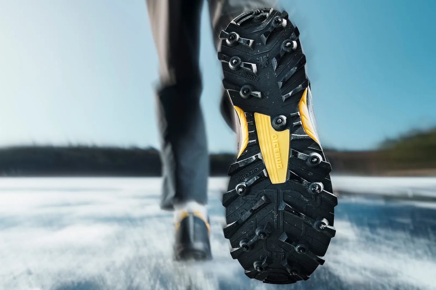 Bottom view of a studded running shoe in motion on ice, showing the “IceBug” logo on the sole.