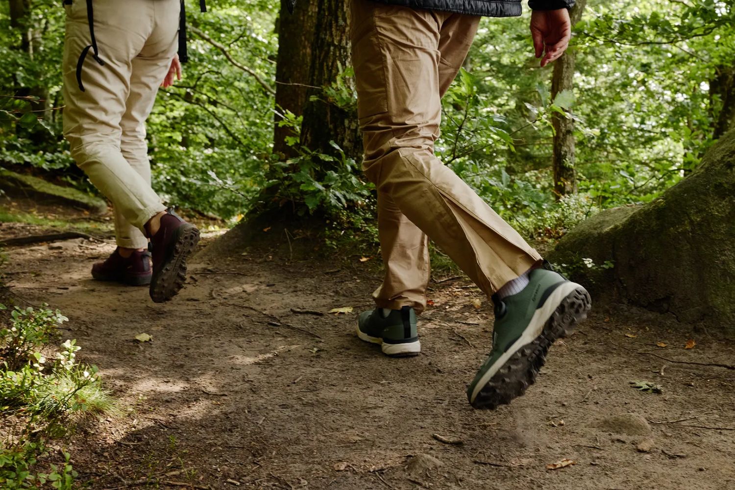 Two people hike on a forest trail, shown from behind with focus on their shoes and rugged path.