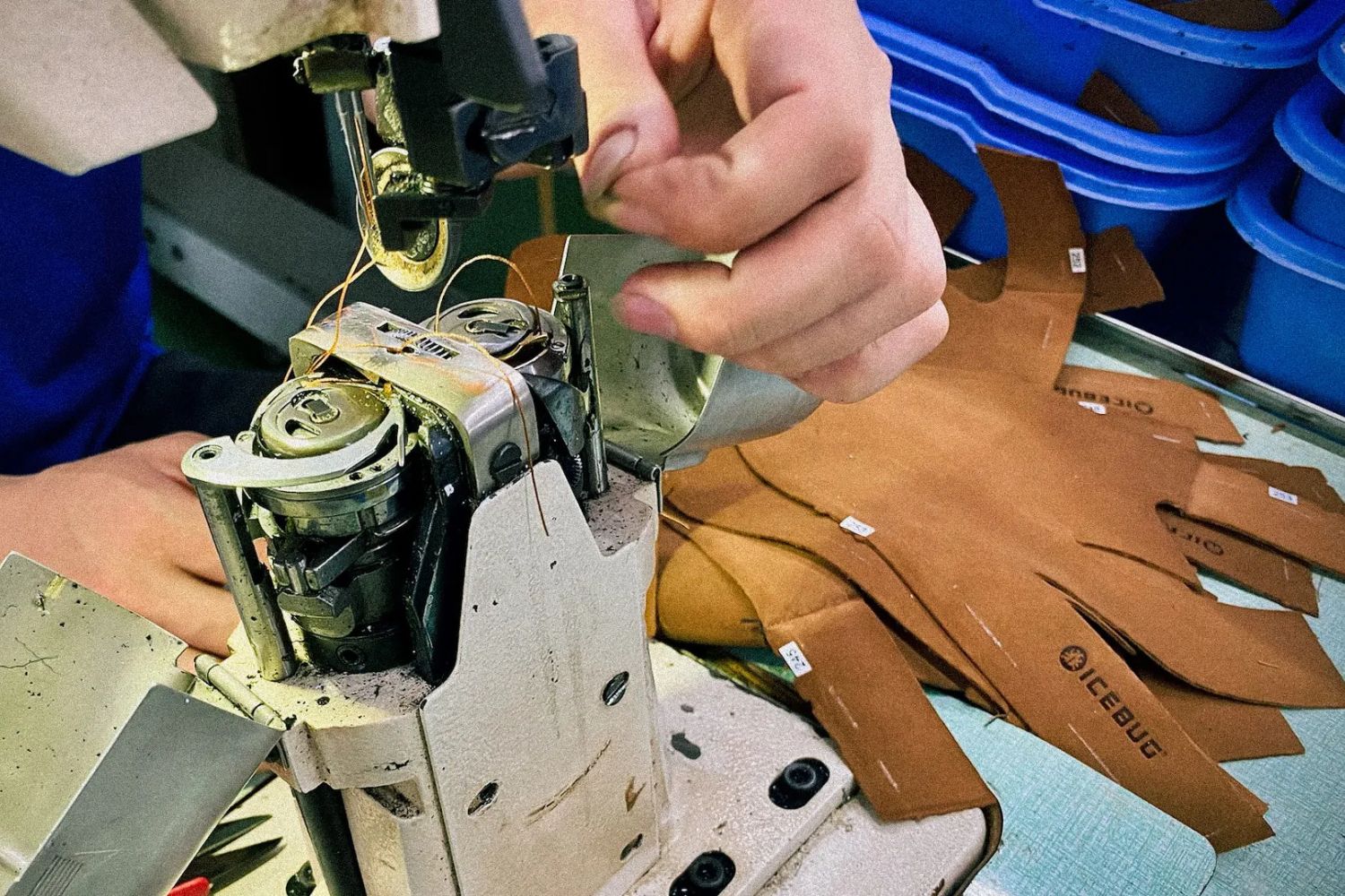 Close-up of hands sewing brown shoe parts labeled “IceBug” on a machine in a workshop.