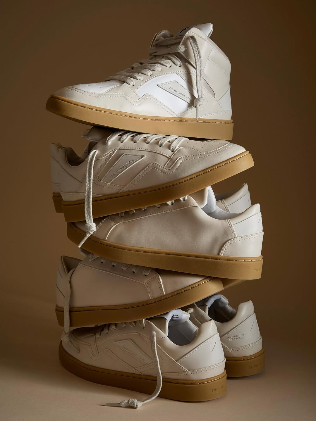 A stacked tower of cream-colored Thousand Fell sneakers, including both low-top and high-top styles, arranged against a warm beige background. The shoes have smooth uppers, tan rubber soles, and visible laces, creating a sculptural, balanced composition.
