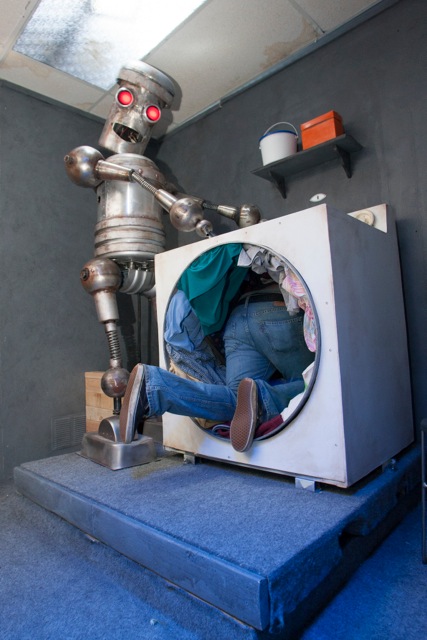 A man crawls though a washing machine as a robot looks on 