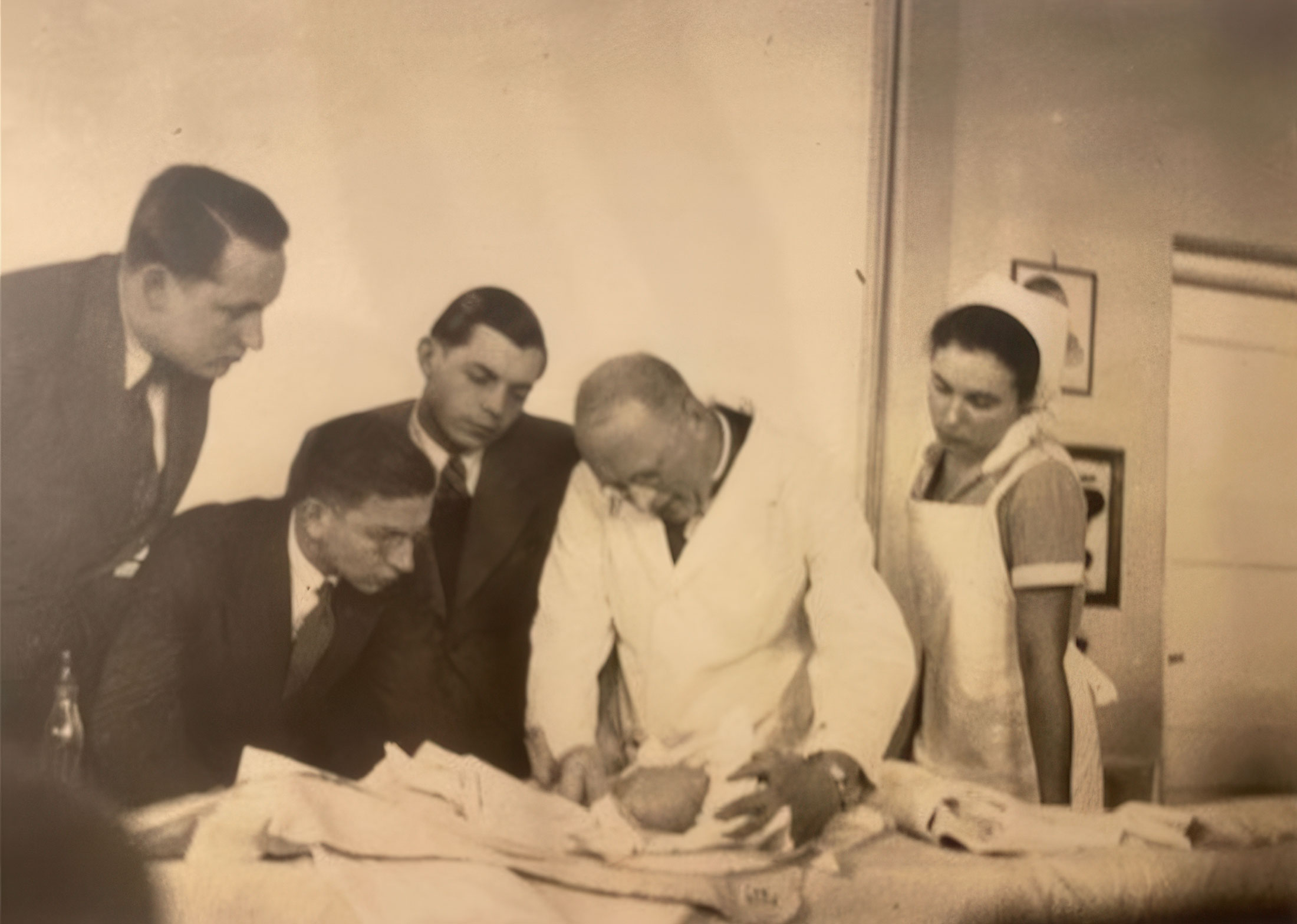 Historische Szene aus der Kinderklinik Würzburg: Ein Arzt untersucht ein Baby, umgeben von Studenten und einer Krankenschwester.