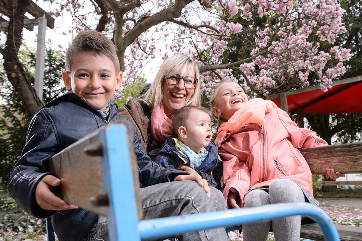 Eine Junge Frau mit drei Kindern auf einem Spielgerät im Freien unter einem blühenden Magnolienbaum, alle lachen.