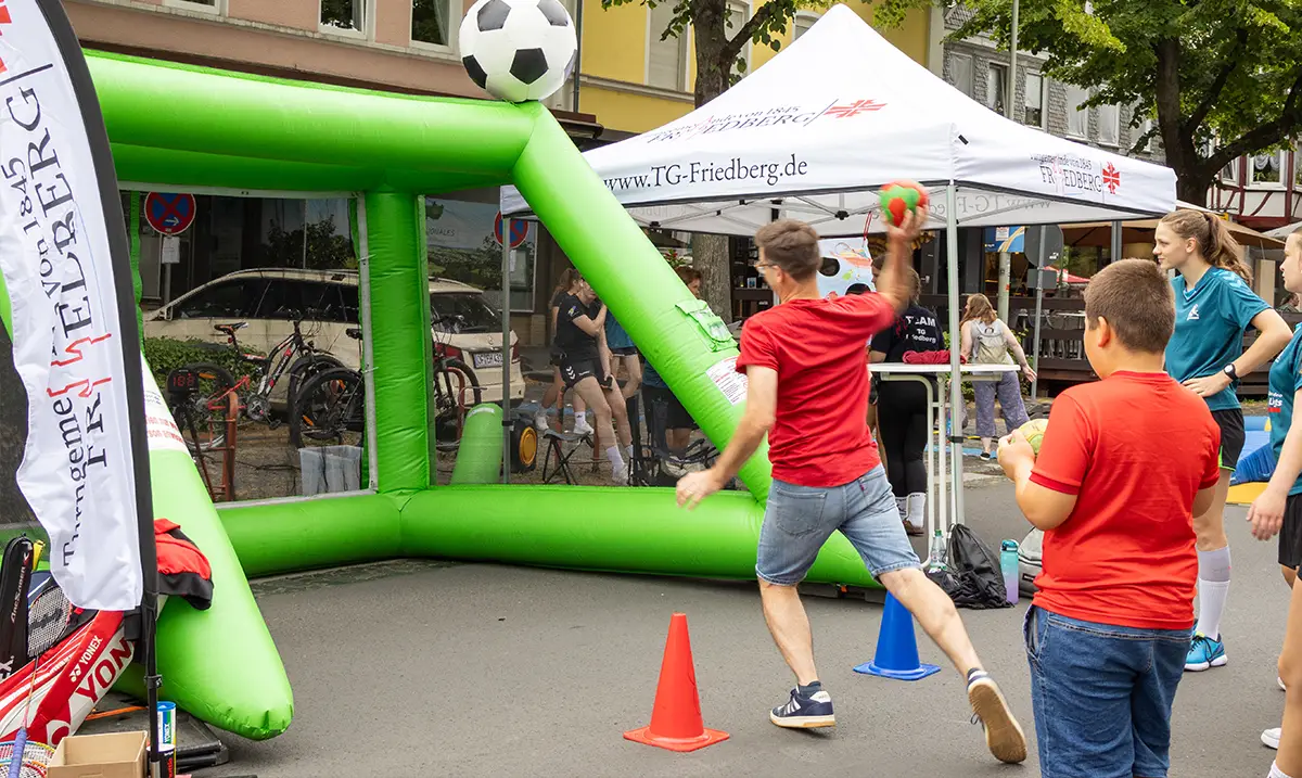 Ein Mann steht auf der Straße vor einem großen grünen Handballtor und wirft einen Ball hinein