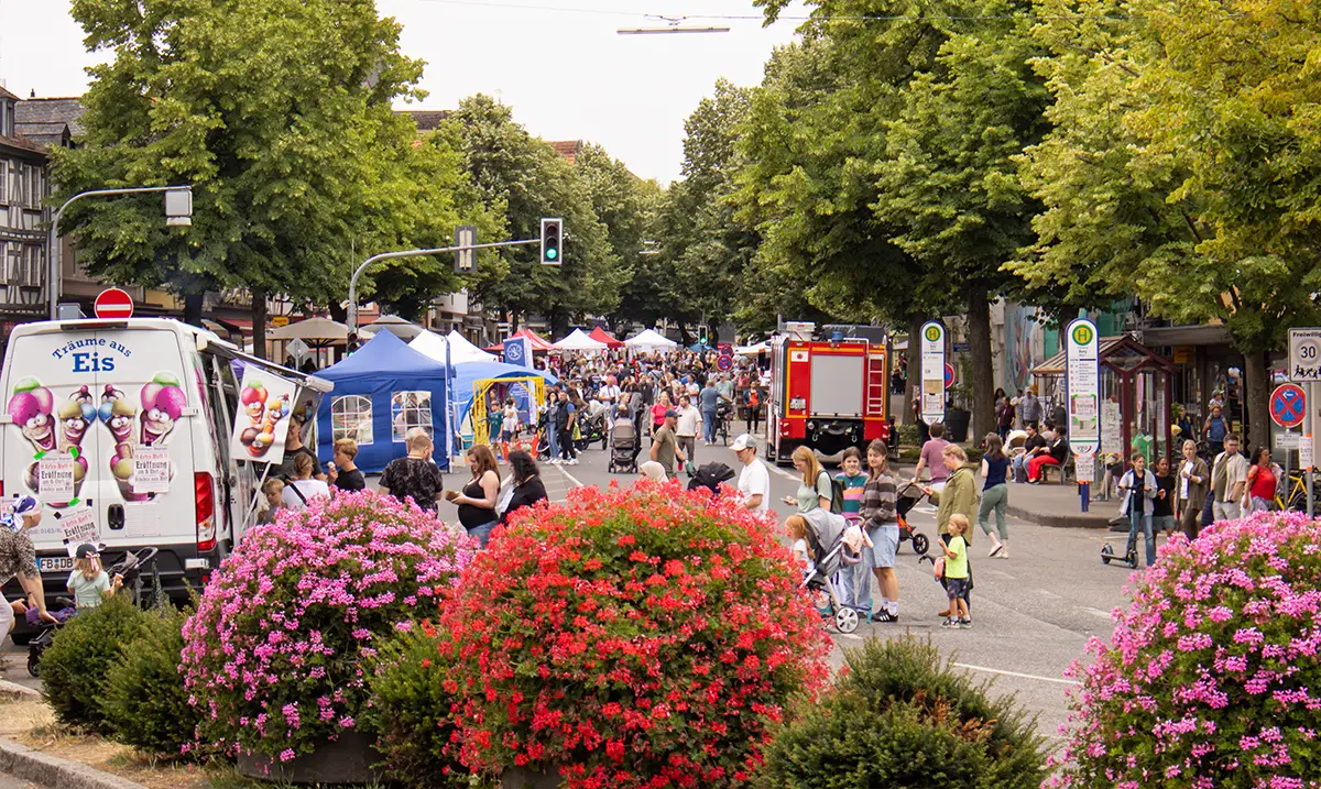 Blühende Sträucher im Vordergrund; im Hintergrund eine Straße mit Menschen und Zelten