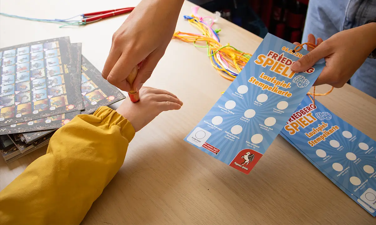 Eine Person stempelt die Hand eines Kindes und gibt ihm eine Karte auf der steht Friedberg Spielt Laufspiel-Stempelkarte