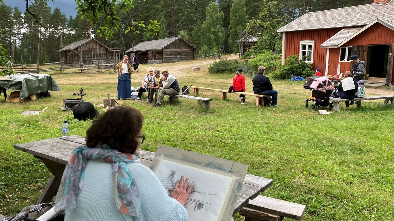Nainen piirtää hiilellä puun alla, takana ihmisiä,