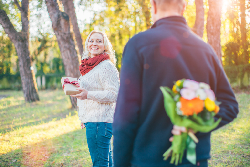 Älterer Mann versteckt Blumenstrauß hinter dem Rücken, sie mit Geschenk in der Hand Älterer Mann versteckt Blumenstrauß hinter dem Rücken, sie mit Geschenk in der Hand