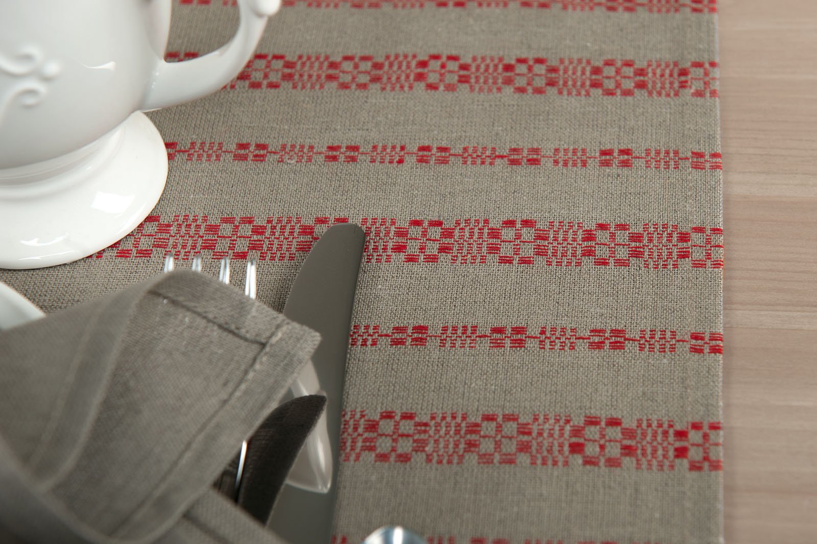 Pilkos spalvos puslininį (lino – medvilnės stalo takelį puošia įaustos tautinių motyvų įkvėptos raudonos juostelės.