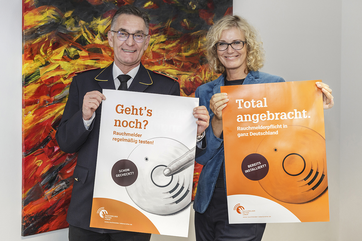 Hartmut Ziebs, Präsident des Deutschen Feuerwehrverbandes (DFV), und Claudia Groetschel, Sprecherin der Initiative "Rauchmelder retten Leben" stellen die Plakate zum Rauchmeldertag 2017 vor. (obs/Rauchmelder retten Leben/Jo Neander)