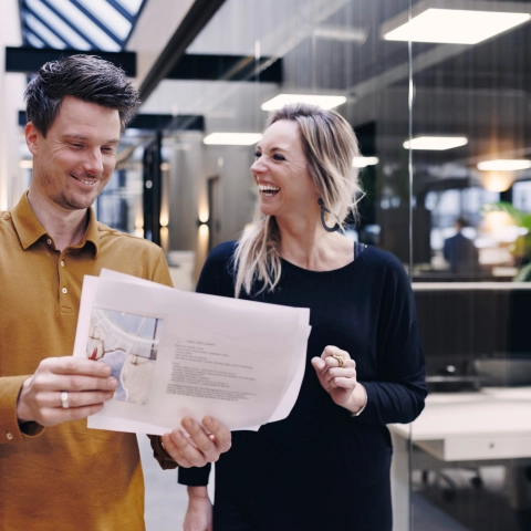 Twee medewerkers die over de gang lopen en enthousiast informatie lezen van een stuk papier af.
