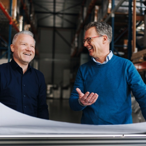 Twee mannen die lachend in gesprek zijn en bij een plotter in het magazijn staan.