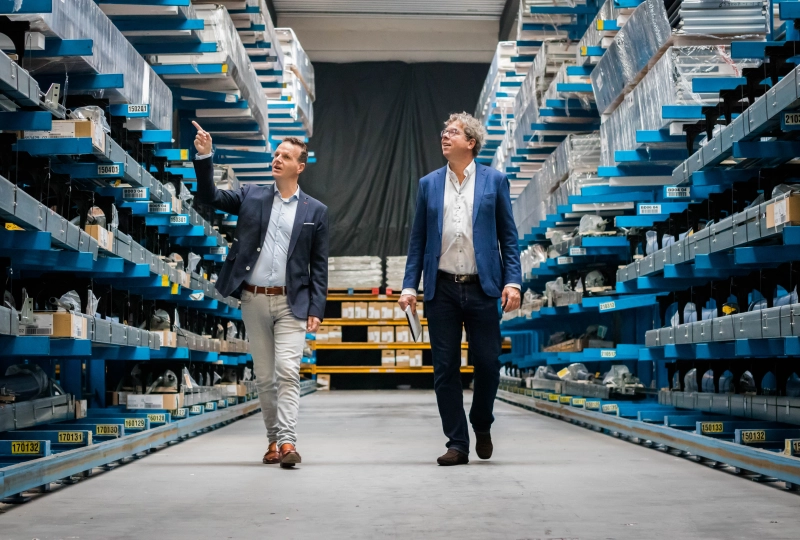Twee mannen lopen door een gangpad van een magazijn waarvan één persoon wijst naar onderdelen die hier liggen.