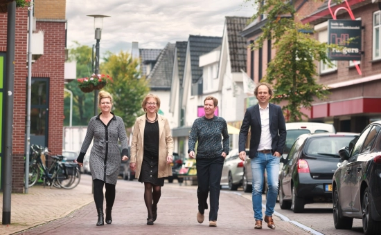 Drie vrouwen en een man lopen naast elkaar op straat, richting de camera. Aan beide kanten staan gebouwen en zijn auto's geparkeerd.