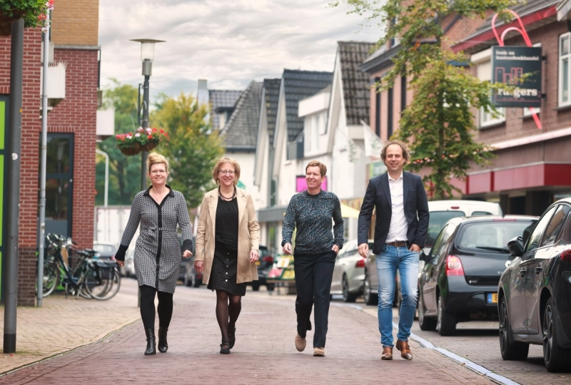 Drie vrouwen en een man lopen naast elkaar op straat, richting de camera. Aan beide kanten staan gebouwen en zijn auto's geparkeerd.