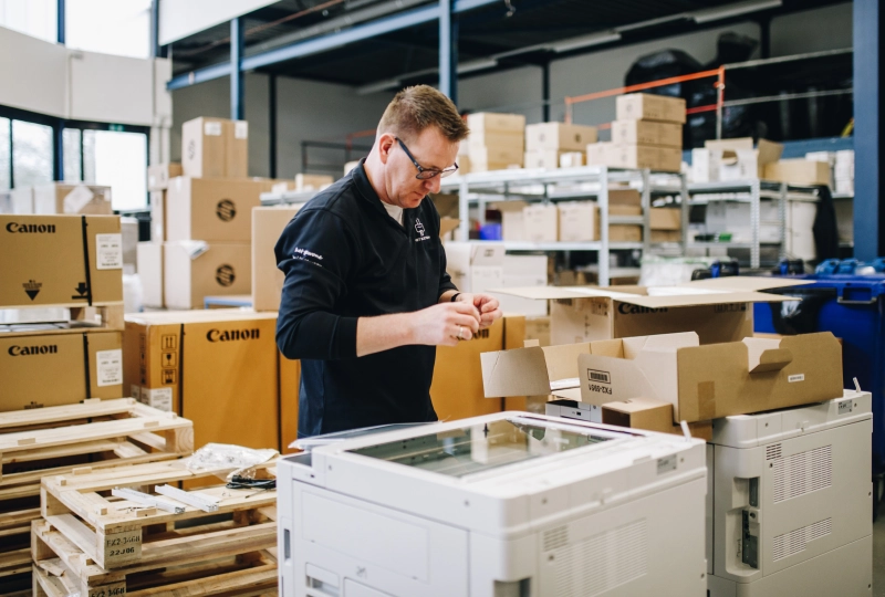 Installateur verricht werkzaamheden aan een printer in een magazijn met veel dozen en materialen op de achtergrond.