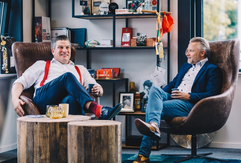 Twee mannen die in een luie stoel zitten op een kantoor en lijken een luchtig gesprek te voeren.