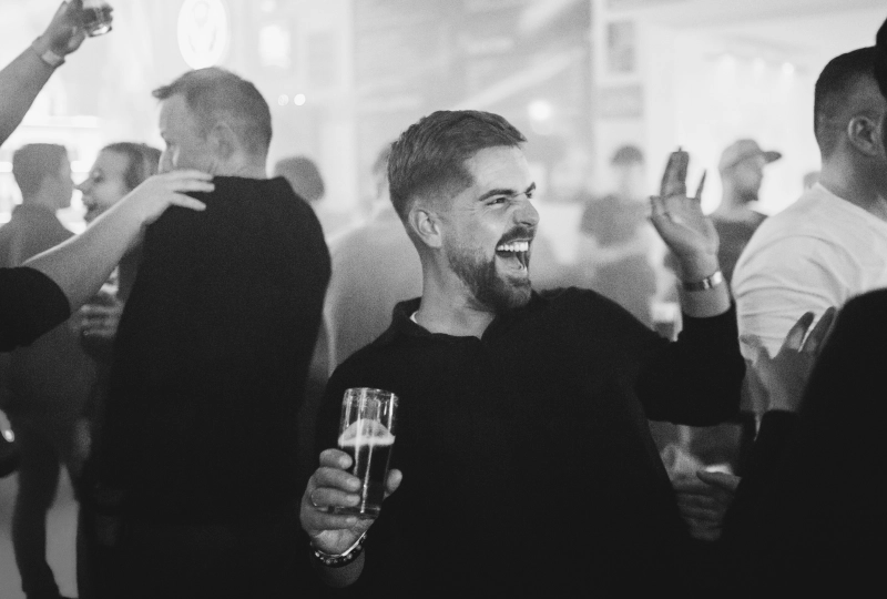 Een man op een feest die uitbundig moet lachen met een drankje in zijn hand en op het punt staat om iemand een high-five te geven.