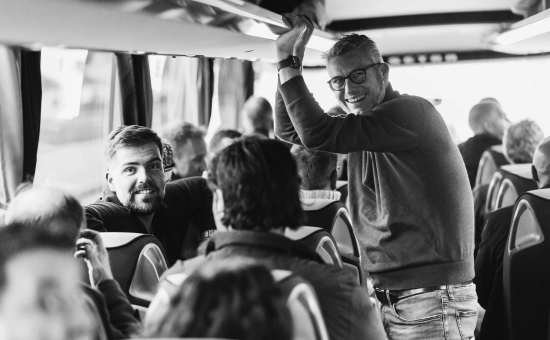 Meerder personen die zitten in een bus waarvan er één iemand staat en samen met iemand die zit, lachend in de camera kijken.