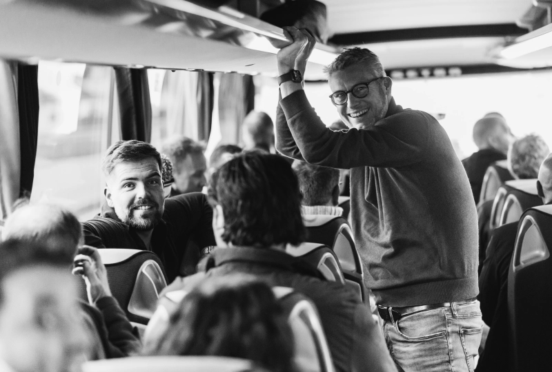 Meerder personen die zitten in een bus waarvan er één iemand staat en samen met iemand die zit, lachend in de camera kijken.