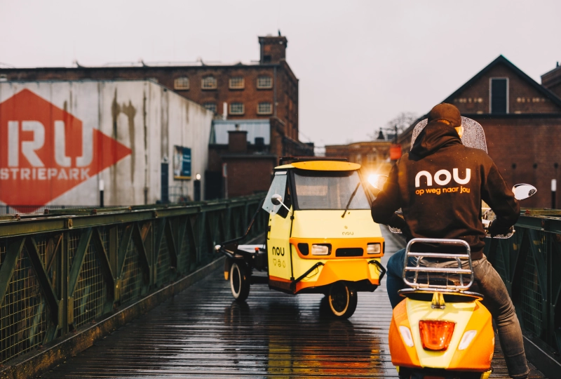 Een persoon zittend op een scooter die staat op een kleine brug. Daarbij staat er ook een tuktuk op de brug in een avondsfeer.