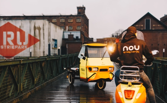 Een persoon zittend op een scooter die staat op een kleine brug. Daarbij staat er ook een tuktuk op de brug in een avondsfeer.