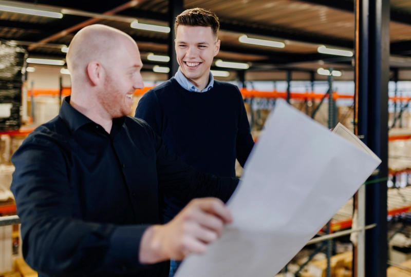 Twee mannen die lachend kijken naar een A3-formaat stuk papier in een magazijn ruimte.