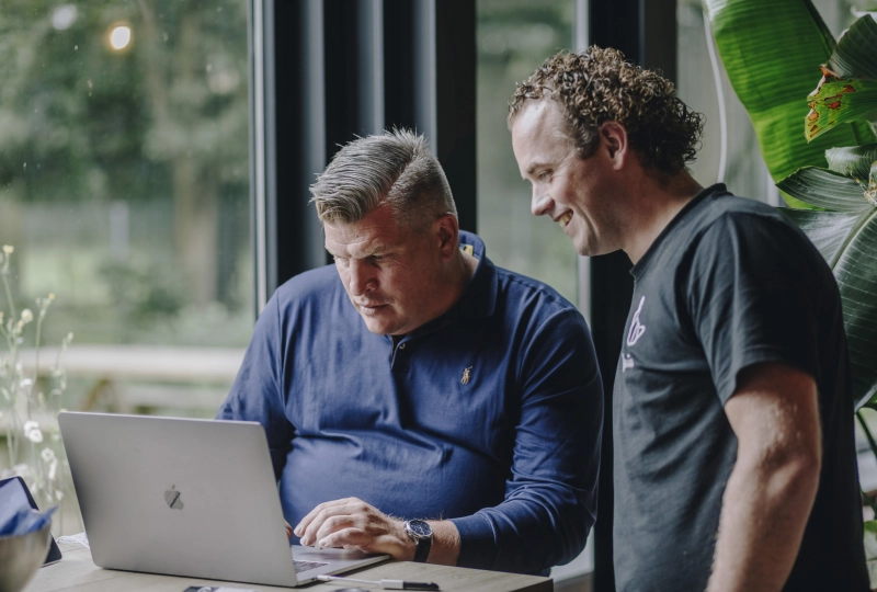 Twee mannen waarvan er één heel moeilijk naar een laptopscherm kijkt en de andere man lachend kijkt.