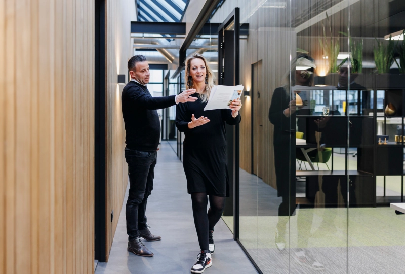 Zakelijke bespreking in moderne kantooromgeving: twee collega's bespreken een document in een lichte, stijlvolle gang met glazen wanden en houten panelen.