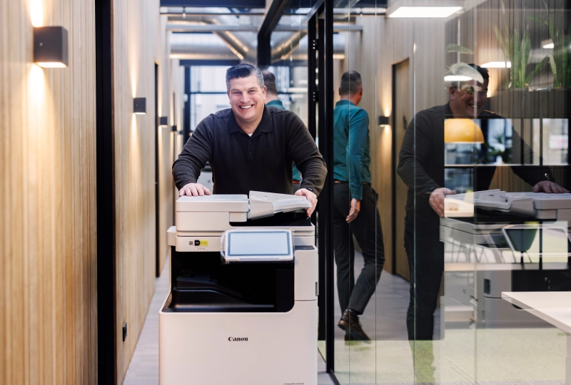 Persoon die enthousiast een printer vooruit duwt over de gang van een kantoor.