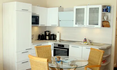 Wohnküche mit Kühlschrank, Mikrowelle, Ceran-Kochfeld, Backofen, Geschirrspüler sowie Esstisch und Rattan-Stühlen.