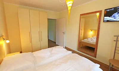 Schlafzimmer mit Doppelbett (1,80 m x 1,90 m), Nachttischkonsolen, Smart-TV, geräumiger Kleiderschrank, großer Wandspiegel und einem Herrendiener mit Sitzmöglichkeit