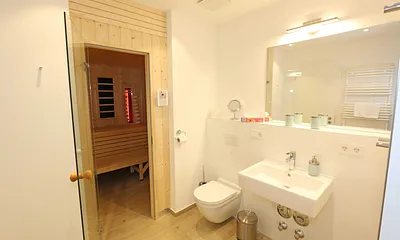 innenliegendes Badezimmer mit Waschtisch, Wandspiegel, ebenerdige Regendusche, WC, Fön, Handtuchtrockner, Kosmetikspiegel und Infrarotsauna