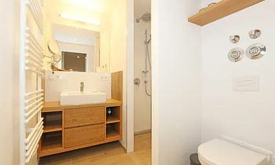 Badezimmer mit einer ebenerdige Regendusche, Handtuchtrockner, Waschtisch mit Schubladen und Ablagefächern, Wandspiegel, WC, Fön und Kosmetikspiegeln