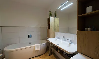 Badezimmer mit ebenerdiger, komfortabler Regendusche mit Echtglasabtrennung, Fön, Kosmetikspiegel, Waschplatz mit Unterschrank sowie freistehender Badewanne