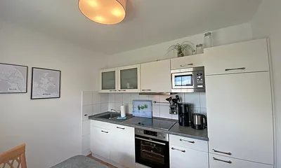 angrenzende Küchenzeile mit Geschirrspüler, Mikrowelle, Ceranfeld, Backofen, Kühlschrank mit Gefrierfach, Wasserkocher, Toaster, Kaffeemaschine und diverse Küchenutensilien