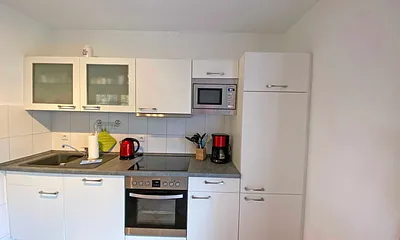 angrenzende Küchenzeile mit Geschirrspüler, Mikrowelle, 4 Zonen-Ceranfeld, Backofen, Kühlschrank mit Gefrierfach, Wasserkocher, Toaster, Kaffeemaschine und diversen Küchenutensilien 