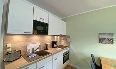 offene Küchenzeile mit Ceranfeld, Kompaktbackofen, Geschirrspüler, Mikrowelle, Spüle, Kühlschrank mit Gefrierfach und diverse Elektrokleingeräte und Küchenutensilien