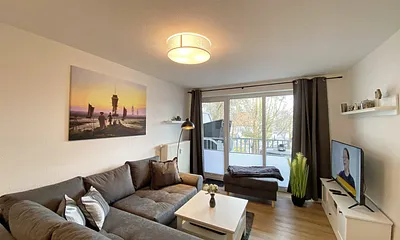 Wohnbereich mit gemütlicher Couch mit Ottomane, Couchtisch, Leselampe, kleines Highboard sowie TV-Board mit Smart-TV und DAB-Radio mit Bluetooth, USB und AUX