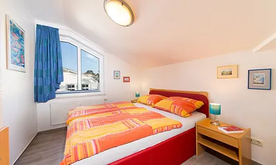 Das Schlafzimmer mit gemütlicher Dachschräge und Doppelbett sowie Gardine und Plissee am Fenster. 