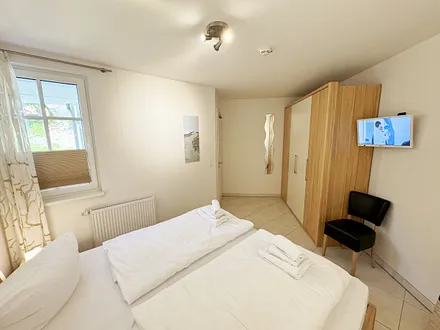 Schlafzimmer mit komfortablem Doppelbett (1,80 m x 2,00 m), hochwertigen Matratzen, Nachttischkonsolen, 4-trg. Drehtürenkleiderschrank (Kernbuche mit Lack creme Fronten), Verdunklungsplissees & TV