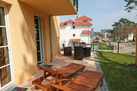 Eckterrasse (Süd/Ost) mit 2 Teakholzliegen und ein Ensemble mit Tisch und vier Stühlen und einem Blick auf die Ostsee durch lichte Baumkronen hindurch