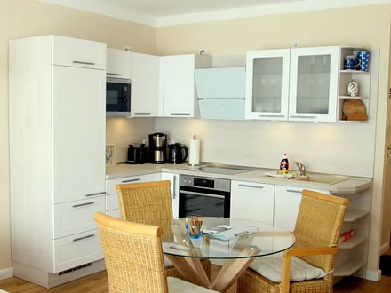 Wohnküche mit Kühlschrank, Mikrowelle, Ceran-Kochfeld, Backofen, Geschirrspüler sowie Esstisch und Rattan-Stühlen.