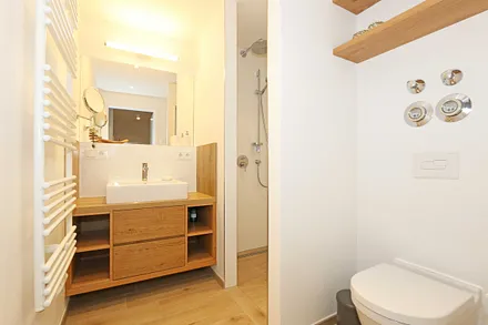 Badezimmer mit einer ebenerdige Regendusche, Handtuchtrockner, Waschtisch mit Schubladen und Ablagefächern, Wandspiegel, WC, Fön und Kosmetikspiegeln