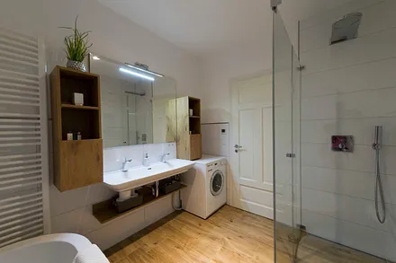 Badezimmer mit ebenerdiger, komfortabler Regendusche mit Echtglasabtrennung, Fön, Kosmetikspiegel, Waschplatz mit Unterschrank sowie freistehender Badewanne