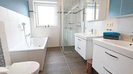 Tageslichtbad mit Badewanne, bodengleicher Echtglasdusche, 2 Waschtische mit Unterschränken und Spiegelschrank, WC, Kosmetikspiegel, Handtuchtrockner, Haarfön