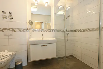 Badezimmer mit ebenerdiger und geräumiger Regen-Dusche, Waschplatz, WC, Fön, Kosmetikspiegel sowie einen Handtuchheizkörper