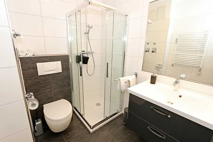 Badezimmer mit Regendusche, Waschtisch, Fön, WC, beleuchteten Spiegel sowie Handtuchheizkörper